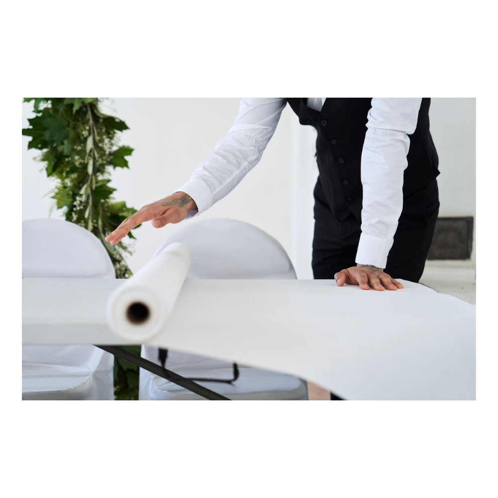 Une personne en tenue de soirée, avec une chemise blanche et un gilet noir, déroule un rouleau de nappe Dunicel (1,18 m x 40 m) de Duni GmbH sur une table ; on aperçoit un peu de vert sur le fond simple.