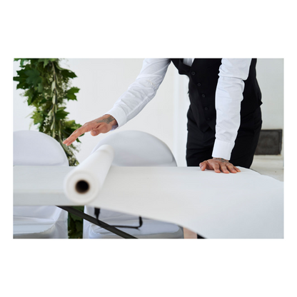 Une personne en tenue de soirée, avec une chemise blanche et un gilet noir, déroule un rouleau de nappe Dunicel (1,18 m x 40 m) de Duni GmbH sur une table ; on aperçoit un peu de vert sur le fond simple.