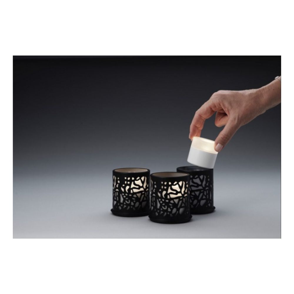 Een hand tilt een kleine witte LED-kaars uit een zwarte houder, terwijl twee identieke Duni GmbH Multicolour LED Set, Mini - 8 stuks in soortgelijke houders op een grijs oppervlak voor een donkere achtergrond rusten.