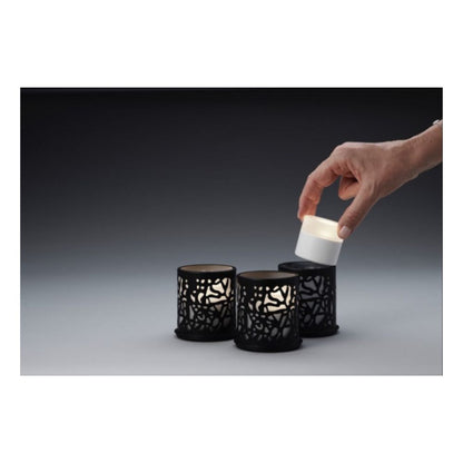 Een hand tilt een kleine witte LED-kaars uit een zwarte houder, terwijl twee identieke Duni GmbH Multicolour LED Set, Mini - 8 stuks in soortgelijke houders op een grijs oppervlak voor een donkere achtergrond rusten.