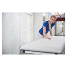 Eine Frau in blauem Hemd und weißer Hose reinigt einen weißen Untersuchungstisch mit den Bacillola® Zero Tissues Schnell-Desinfektionstüchern (1 Stück) der Paul Hartmann AG zur umweltfreundlichen Desinfektion in einer hellen medizinischen Umgebung.