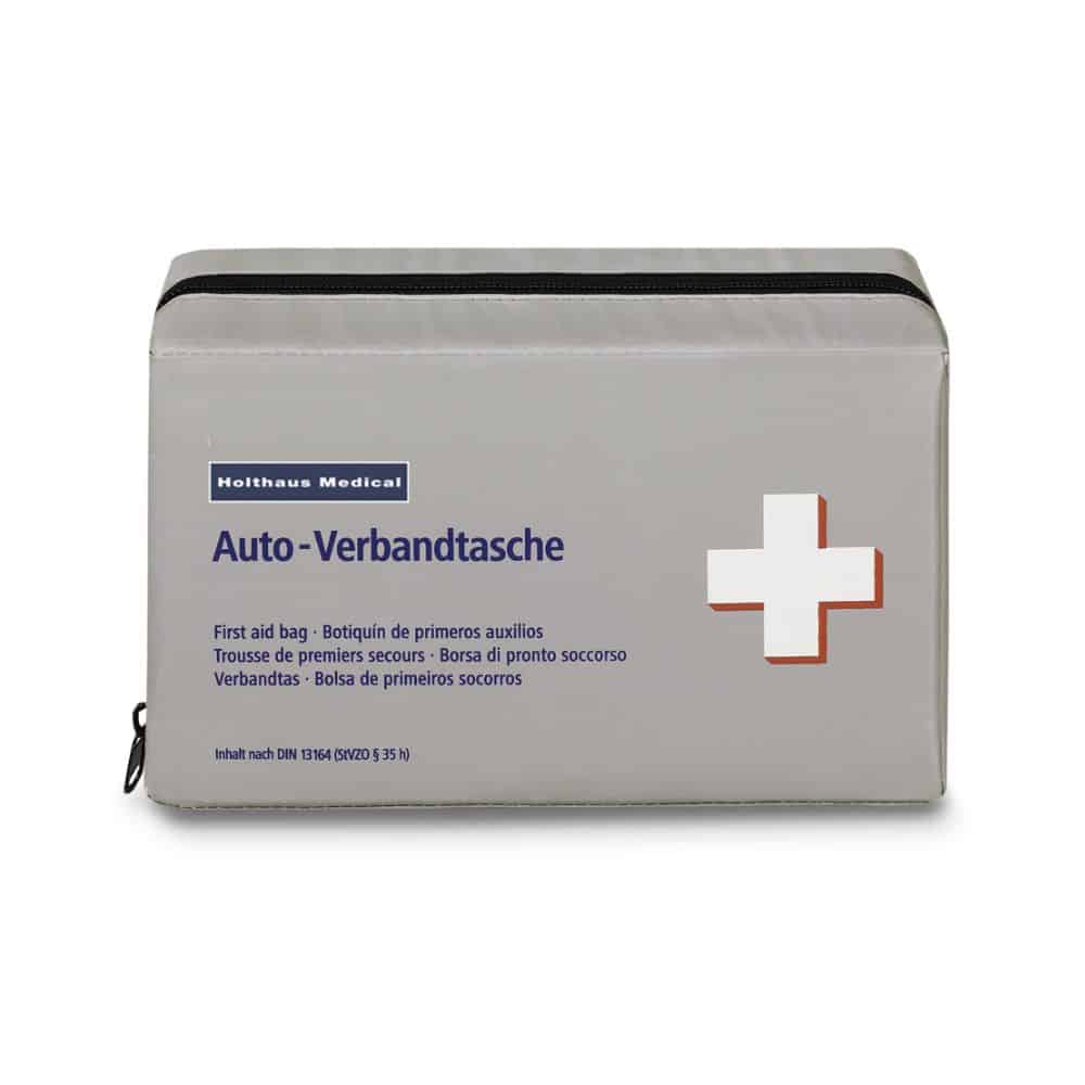 Een rechthoekige, grijze verbandtas met zwarte ritssluiting. De tas draagt de tekst "Holthaus Medical Klassik Verbandtas" en "Auto-Verbands-Tasche" en heeft rechts een wit kruis met een rode rand. Bovenaan is het gemarkeerd met het merk "Holthaus Medical GmbH & Co. KG", dat voldoet aan DIN 13164.