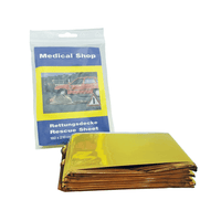 Devant celle-ci se trouve une couverture de survie pliée, de couleur jaune doré, en film polyester recouvert d'aluminium. Derrière celle-ci se trouve un paquet portant l'inscription « Holthaus Medical Rettungsdecke, gold silber » de la société Holthaus Medical GmbH & Co. KG, qui représente une scène d'accident avec une voiture rouge et dont les dimensions sont de 160 x 210 cm.