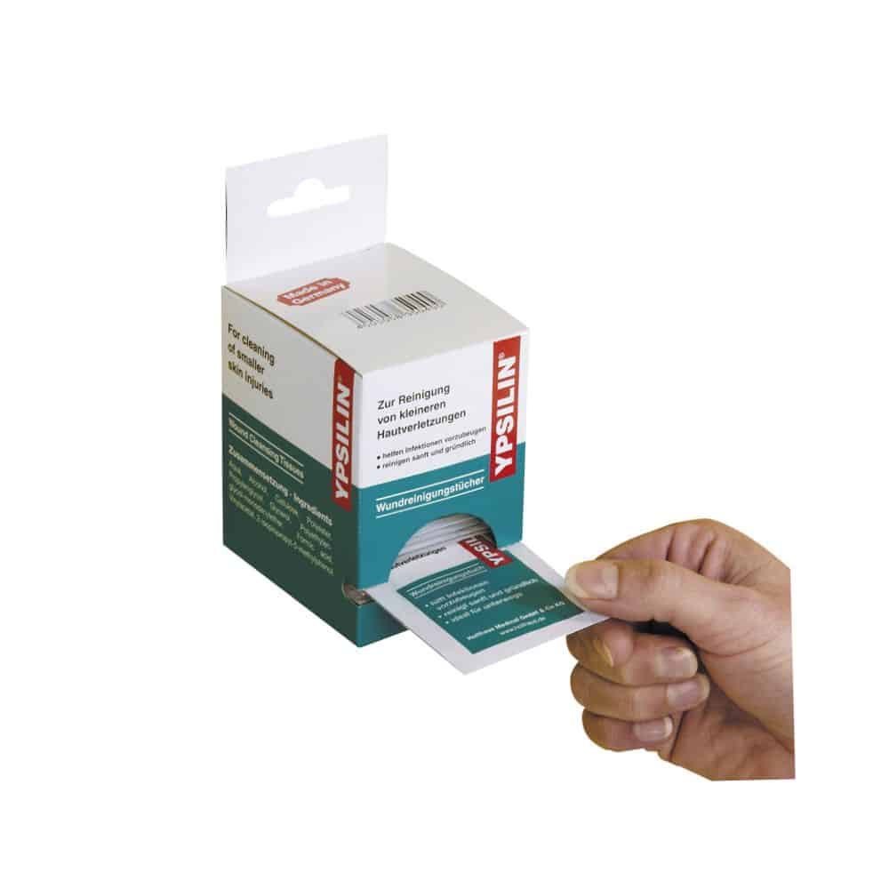Une main tire une lingette de nettoyage de plaies Holthaus Medical YPSILIN® d'un distributeur en carton. L'emballage porte un texte en allemand et la boîte est blanche et verte, avec des informations sur le produit et le nom de la marque Holthaus Medical GmbH & Co. KG bien visibles.