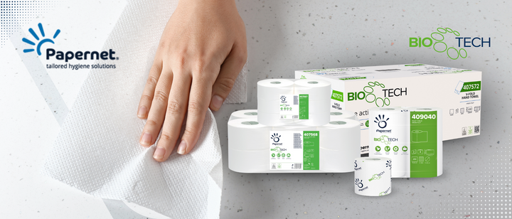 Eine Hand wischt mit einem Papierhandtuch eine weiße Fläche ab. Auf der rechten Seite sind mehrere Verpackungen von Papernet Bio Tech Toilettenpapier ausgestellt, die das Produktbranding und die grünen umweltfreundlichen Etiketten zeigen.