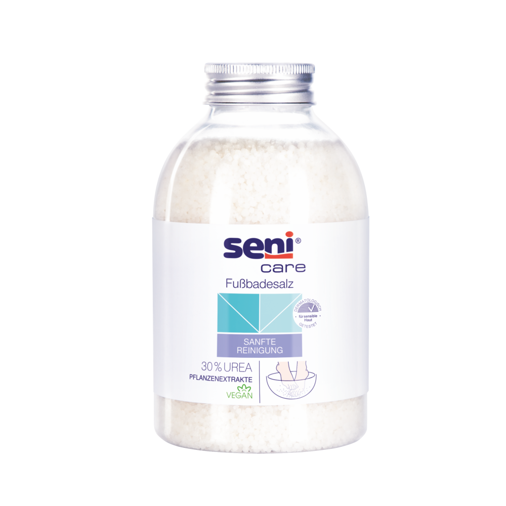 Een heldere pot met een zilveren deksel bevat witte uitloopartikelen: Seni Care badzout voor ontspannende voetbaden (400g) van TZMO Deutschland GmbH. Op het etiket staat 30% ureum, veganistische formule, zachte reiniging en plantenextracten voor ontspannende voetbaden.
