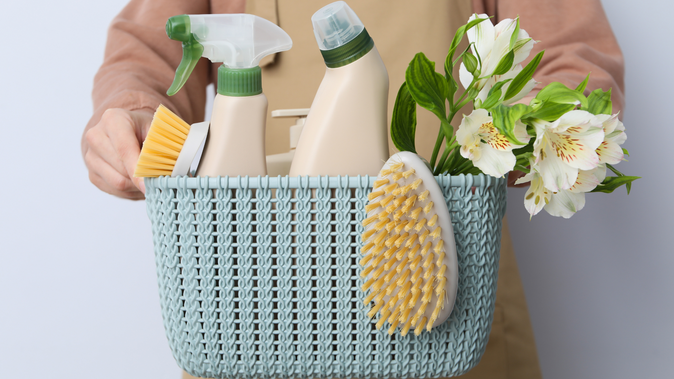 Eine Person hält einen hellblauen geflochtenen Korb mit Reinigungsmitteln, einer Bürste mit gelben Borsten, einer Sprühflasche, einer Plastikflasche, einem Schrubber und einem kleinen Strauß weißer Blumen.