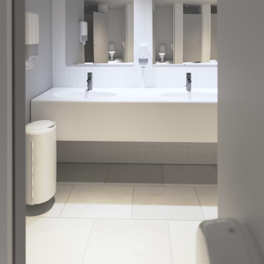 Une salle de bain propre et moderne présente un design Elevation avec un long plan de travail blanc sur lequel se trouvent deux lavabos avec des robinets chromés. Les murs sont ornés de carreaux blancs et des miroirs sont installés au-dessus des lavabos. Les distributeurs Tork 561500 pour savon et désinfectant pour les mains Elevation S4 de TORK – Essity Professional Hygiene Germany GmbH sont élégamment montés au mur. Une élégante poubelle blanche se trouve sur le sol.