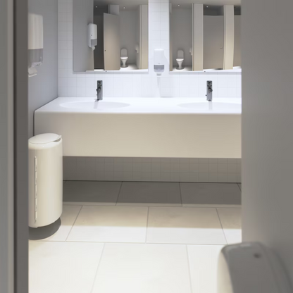 Une salle de bain propre et moderne présente un design Elevation avec un long plan de travail blanc sur lequel se trouvent deux lavabos avec des robinets chromés. Les murs sont ornés de carreaux blancs et des miroirs sont installés au-dessus des lavabos. Les distributeurs Tork 561500 pour savon et désinfectant pour les mains Elevation S4 de TORK – Essity Professional Hygiene Germany GmbH sont élégamment montés au mur. Une élégante poubelle blanche se trouve sur le sol.