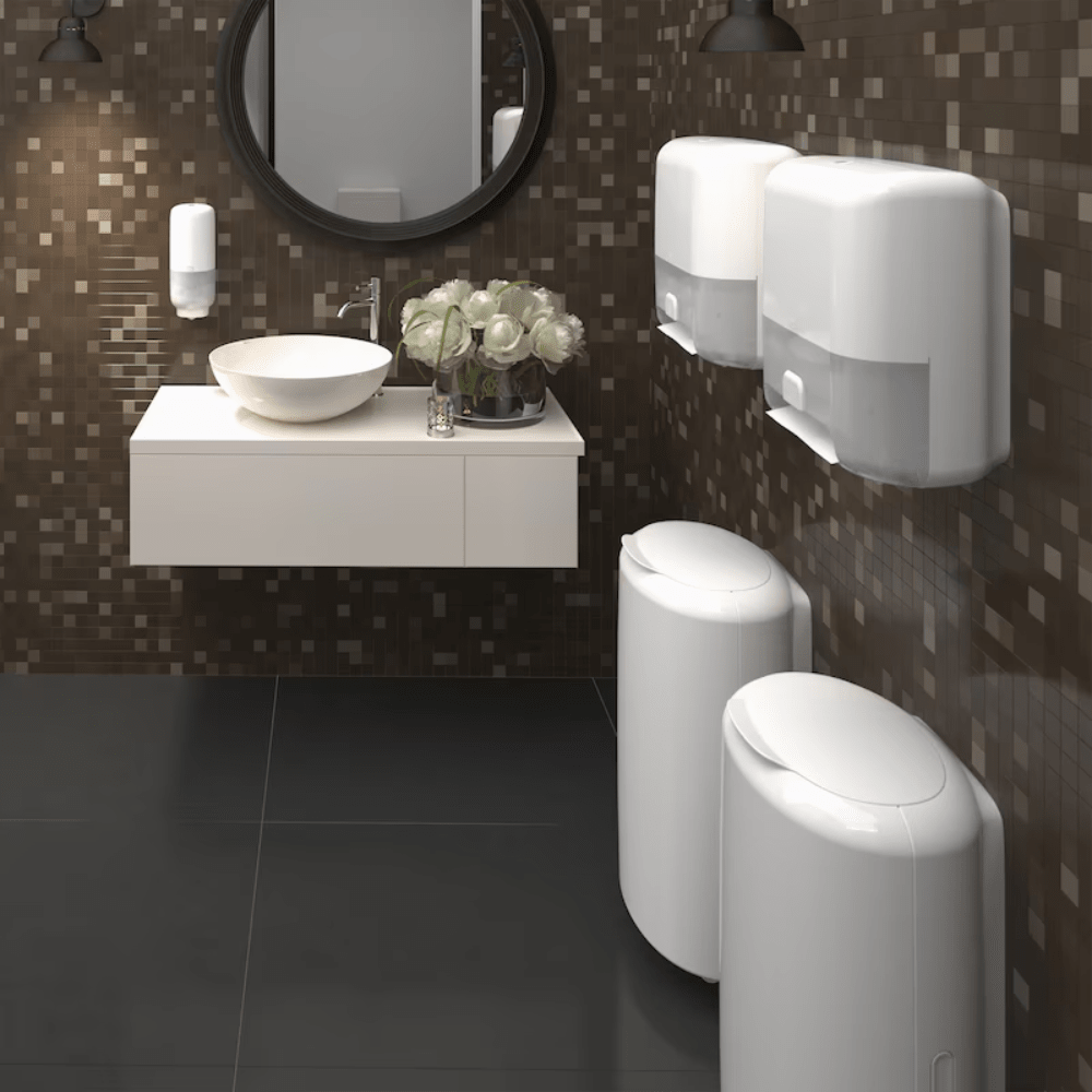 Een moderne badkamer met donkere mozaïektegels aan de muren en een zwarte tegelvloer heeft een ronde spiegel boven een wandgemonteerde wastafel met een wit opzetbekken. Twee Tork 561600 huidverzorgingsdispensers met Intuition Sensor van TORK – Essity Professional Hygiene Germany GmbH zorgen voor contactloze uitgifte, en rechts staan twee afvalbakken. Een vaas met witte bloemen siert de wastafel.