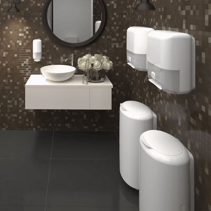 Een moderne badkamer met donkere mozaïektegels aan de muren en een zwarte tegelvloer heeft een ronde spiegel boven een wandgemonteerde wastafel met een wit opzetbekken. Twee Tork 561600 huidverzorgingsdispensers met Intuition Sensor van TORK – Essity Professional Hygiene Germany GmbH zorgen voor contactloze uitgifte, en rechts staan twee afvalbakken. Een vaas met witte bloemen siert de wastafel.