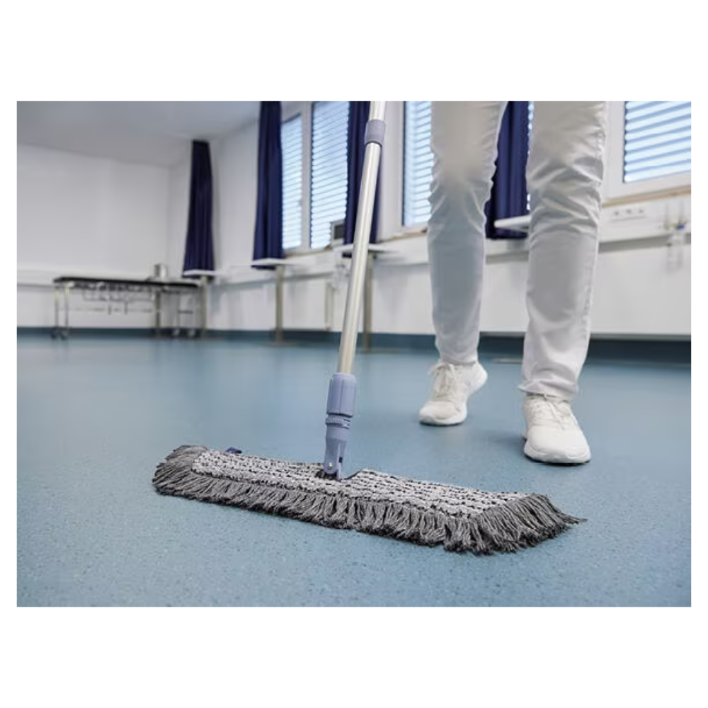 Een persoon in witte schoenen en broek dweilt met een Vileda GmbH Professional Swep Duo r-MicroCombi Mop - 50 cm een blauwe vloer in een kamer met gesloten jaloezieën en blauwe gordijnen. Dichtbij staan een klaptafel en onder de ramen een radiator.