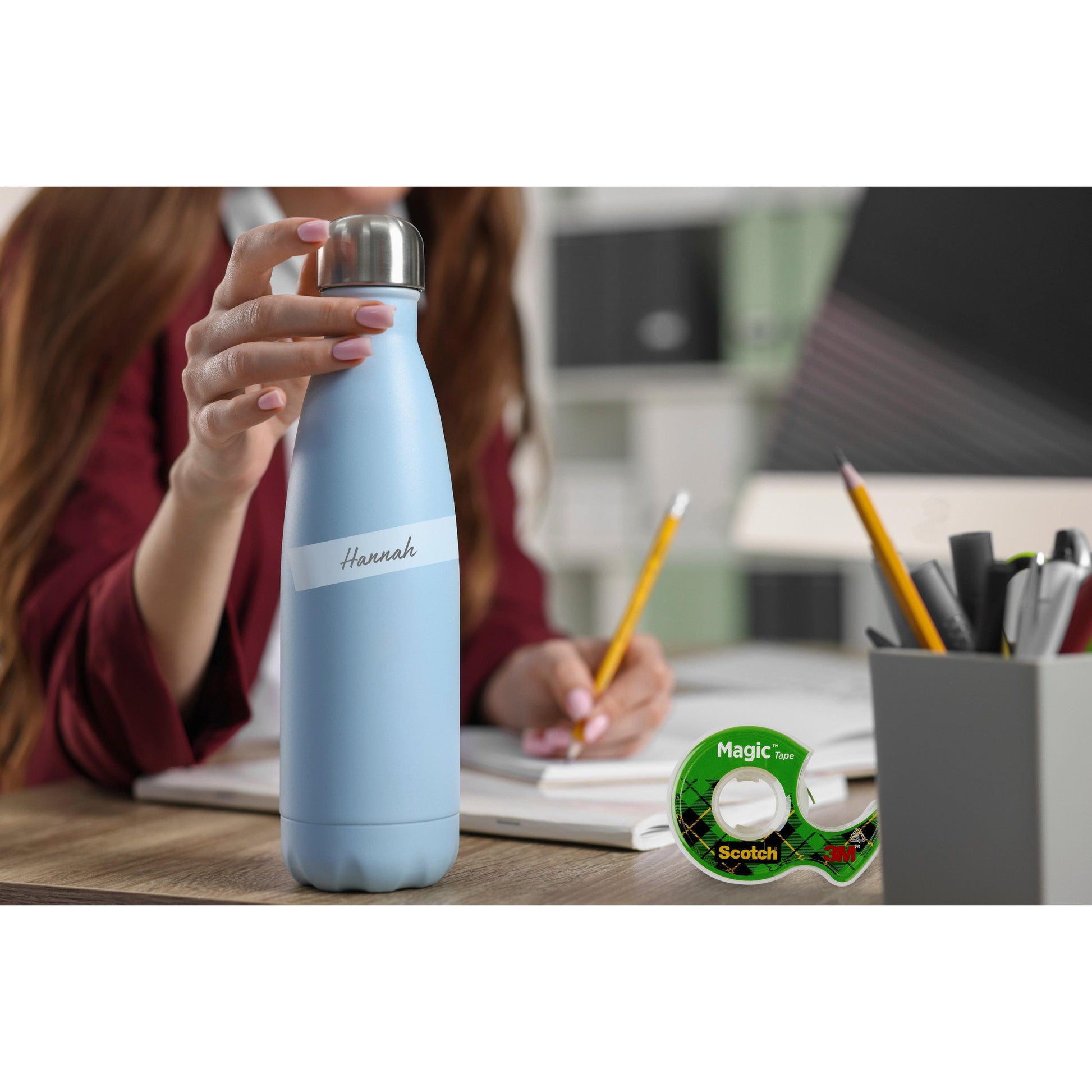 Eine Person an einem Schreibtisch hält eine hellblaue Wasserflasche mit der Aufschrift "Hannah"; daneben liegen Büromaterialien, ein Bleistift und eine Scotch® Magic™ Unsichtbares Klebeband Nachfüllpackung (1 Rolle, 19 mm x 25 m) der 3M Deutschland GmbH.