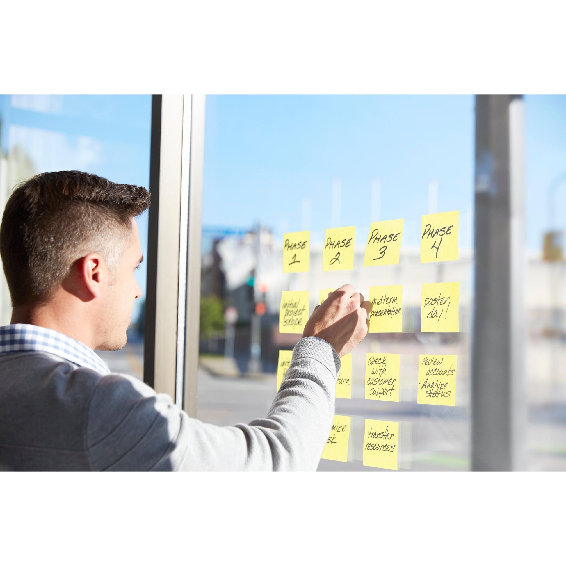 Eine Person in einem hellgrauen Pullover verwendet 3M Deutschland GmbH Post-it® Super Sticky Z-Notes, Gelb (76 × 127 mm), um bei der Planung bei Tageslicht Aufgaben mit den Bezeichnungen "Phase 1" bis "Phase 4" an einem Glasfenster zu organisieren.