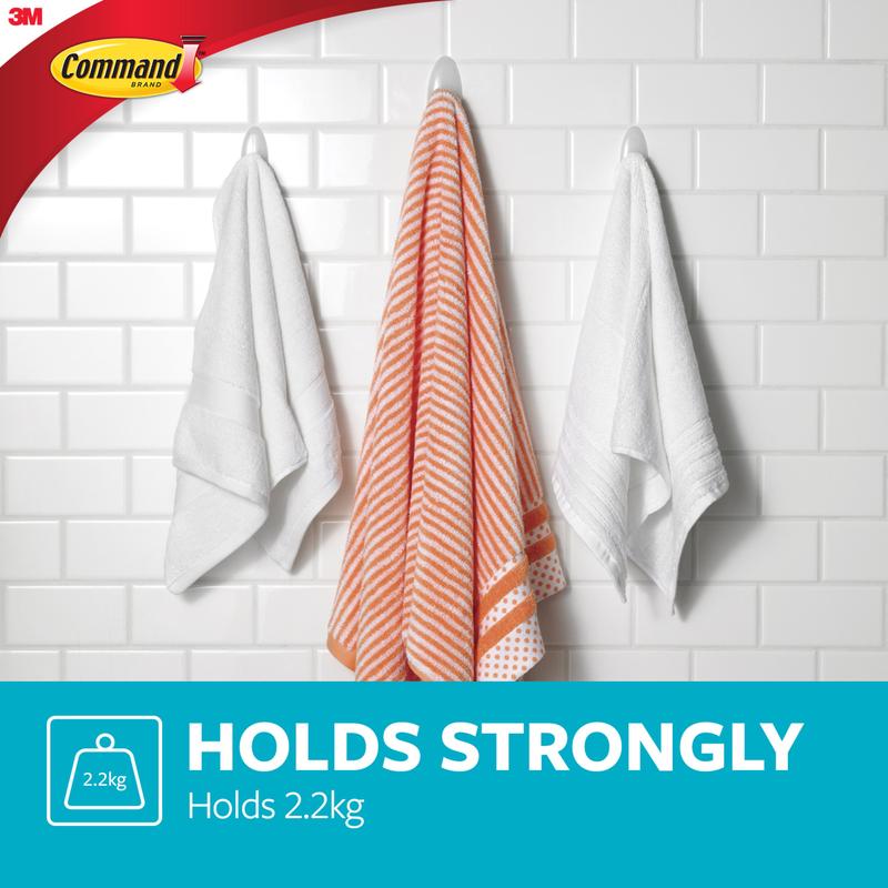 Trois serviettes sont suspendues à des crochets pour serviettes de bain Command™ de 3M Deutschland GmbH - une serviette rayée orange et blanche entre deux blanches - sur un mur carrelé blanc. Texte : "HÄLT STARK Supporte 2,2 kg." Logos Command et 3M visibles.