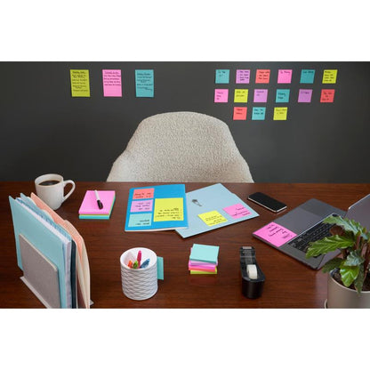 Een opgeruimd bureau met laptop, koffie, pennen en kantoorbenodigdheden staat voor een muur die bedekt is met rijen van 3M Deutschland GmbH Post-it® Super Sticky Notes (47,6 mm x 47,6 mm, 90 vel/Blok, 100% PEFC); een lege stoel staat achter het bureau.