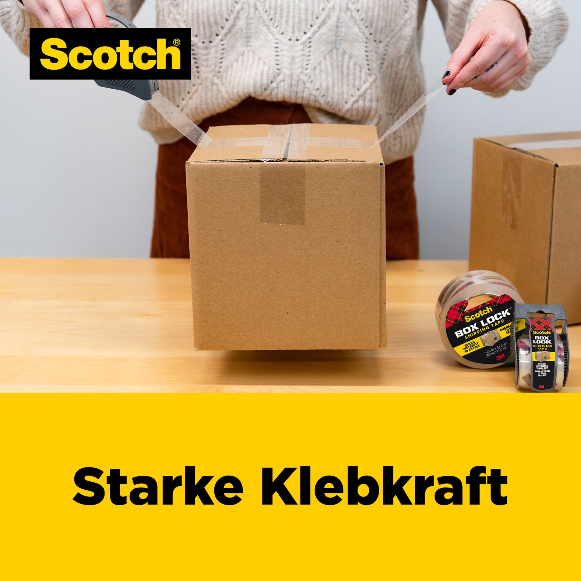 Eine Person verschließt einen Karton mit Scotch® Box Lock™ Verpackungsklebeband 3950-LR3-DC (48 mm x 50 m, 3 Rollen/Packung) der 3M Deutschland GmbH auf einem Holztisch. Das Bild zeigt das Band und "Starke Klebkraft" auf Gelb.