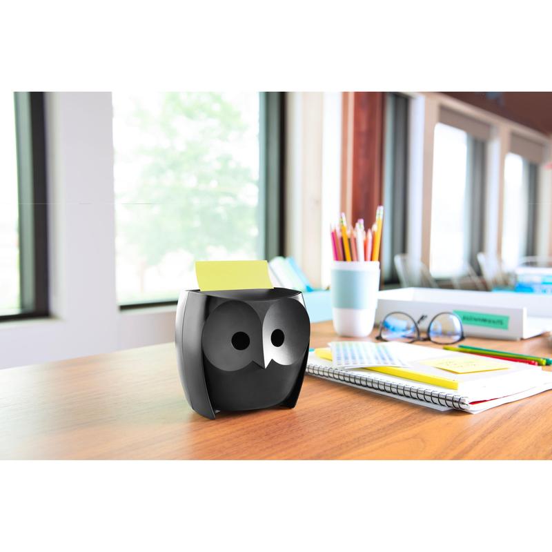 Een Post-it® Z-Notes Dispenser, Uil, Zwart van 3M Deutschland GmbH staat op een houten tafel met notitieboekjes, een kopje, kleurpotloden in een kopje en Post-it® Super Sticky Z-Notes (45 vel/blok, 2 blokken/pak); grote ramen bieden uitzicht op bomen.