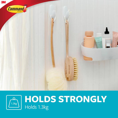 Une salle de bain avec deux crochets pour serviettes de bain Command™ de 3M Deutschland GmbH, qui maintiennent des brosses de bain en bois à côté d'un rideau de douche ; une étagère murale blanche contient des articles de toilette. Texte : "Holds Strongly. Supporte 1,3 kg." Logos Command et 3M visibles.