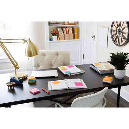 Auf einem schwarzen Schreibtisch stehen ein Laptop, Stifte, eine goldene Lampe, eine Pflanze, Büroartikel und 3M Deutschland GmbH Post-it® Super Sticky Notes 654-6SS-PPNK (rosa, 76x76mm), neben einem beigen Stuhl; Bücher und Kisten füllen die weißen Regale dahinter. Auf der rechten Seite hängt eine Wanduhr.