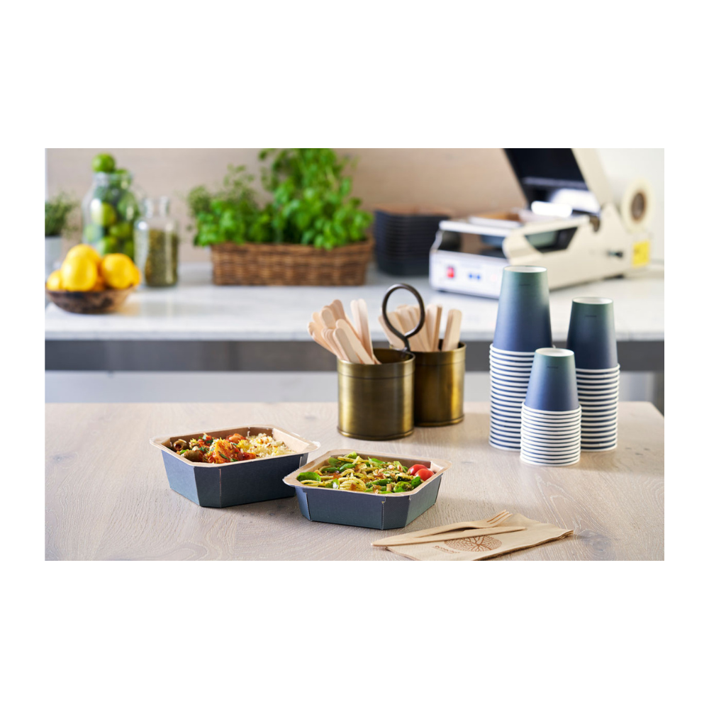 Op de tafel staan twee afhaalkommen, geflankeerd door gestapelde Duni Urban Koffiebekers PAP/PLA van Duni GmbH, houten bestek in een houder en servetten. Citroenen, glazen en kruiden op de achtergrond dragen bij aan de duurzame sfeer van dit eetbereidingsgebied.